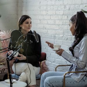 Two women engaged in a podcast interview, sitting on chairs with microphones in front of them, one holding a cup, against a white brick wall background.